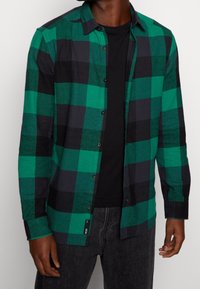 Hombre vistiendo una camisa de cuadros grandes verde y negra sobre una camiseta negra y pantalones vaqueros oscuros, de pie contra un fondo liso.