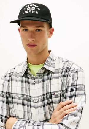 Młody mężczyzna o niebieskich oczach, noszący czarną czapkę Tommy Jeans oraz czarno-białą koszulę w kratę, z założonymi skrzyżowanymi ramionami.