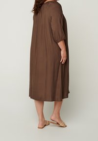 Robe marron mi-longue en tissu lisse avec une coupe décontractée. Présente des manches trois-quarts et un col légèrement froncé.