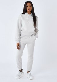 Conjunto de sudadera y pantalones de chándal gris claro en material suave, con un bolsillo frontal, cordones y bordado de logo. Combinado con zapatillas blancas.