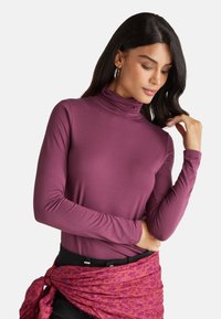 Magenta Langarm-Turtleneck-Shirt aus glattem Stoff, mit taillierter Passform und betontem Kragen, kombiniert mit einem gemusterten pinken Schal.