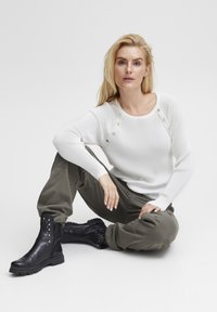 Bijela rebrasta sweatshirt s dekorativnim gumbima na ramenu, uparena s maslinasto zelenim hlačama i crnim kožnim ankle čizmama s zakovicama.
