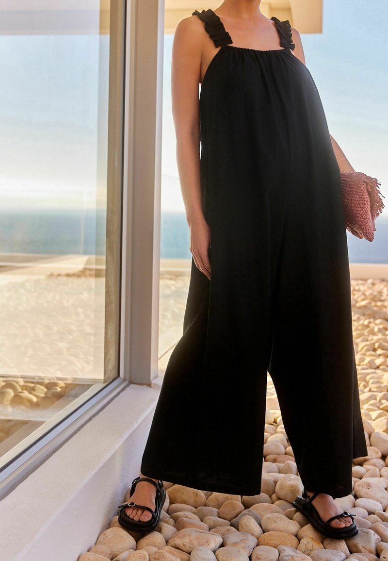 Combinaison noire sans manches avec des bretelles volantées, coupe ample, tissu fluide, assortie à des sandales noires épaisses et à une pochette texturée rose.