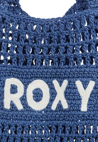 Blauwe gehaakte stof met een open weefsel. Witte "ROXY" letters prominent weergegeven in het midden, met een textuurafwerking.