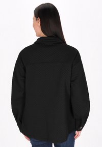 Geacă neagră matlasată, cu guler ascuțit, umeri căzuți și mâneci cu manșete. Partea din spate prezintă un model texturat în formă de diamant.