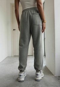 Pantaloni della tuta grigi realizzati in tessuto morbido con vita elasticizzata, caviglie elastiche e una tasca posteriore. Abbinati a scarpe da ginnastica bianche.