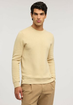 Giovane uomo con capelli castani ricci indossa un maglione beige lavorato e pantaloni marrone chiaro, in piedi davanti a uno sfondo chiaro e uniforme.