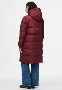 Burgundiškas pūkų paltas su kepure, turintis siuvinėtą dizainą ir ilgą siluetą. Suderintas su plačiomis mėlynomis džinsinėmis kelnėmis ir juodais batais.