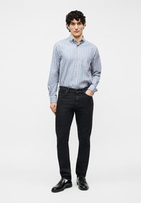 Άνδρας που φοράει μπλε και άσπρο πουκάμισο με ρίγες και κουμπιά, μαύρο τζιν και μαύρα παπούτσια, στέκεται με το ένα χέρι στην τσέπη.