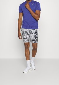 Lila atletiskt t-shirt, korta shorts i kamouflage-mönster i nyanser av grått och svart, vita löparskor och vita ankelsockor.