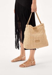 Bézs színű szövött táska, melynek teteje redőzött, aranyszínű láncpántokkal. A texturált minta kiemelkedik a fekete rojtos szoknya és szandálok hátterében.