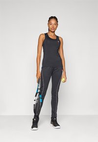 Svart trenings tank topp og bukser, med rosa aksentstriper. Modellen holder en tenniskølle og ball, og viser frem en sporty look.