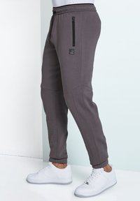 Gray tapered joggers with an elastic waistband, zippered side pocket, and ribbed cuffs, paired with white sneakers on a reflective surface.