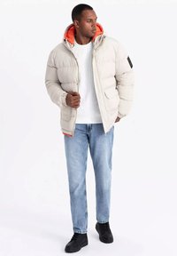 Puffjacka i ljus beige med en orange huva, dragkedja fram och två sidofickor. Bärs över en vit strukturerad sweatshirt och jeans.