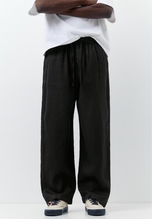 Personne portant un pantalon noir ample à cordon, des baskets blanches avec des semelles noires et une chemise blanche avec les bras croisés.