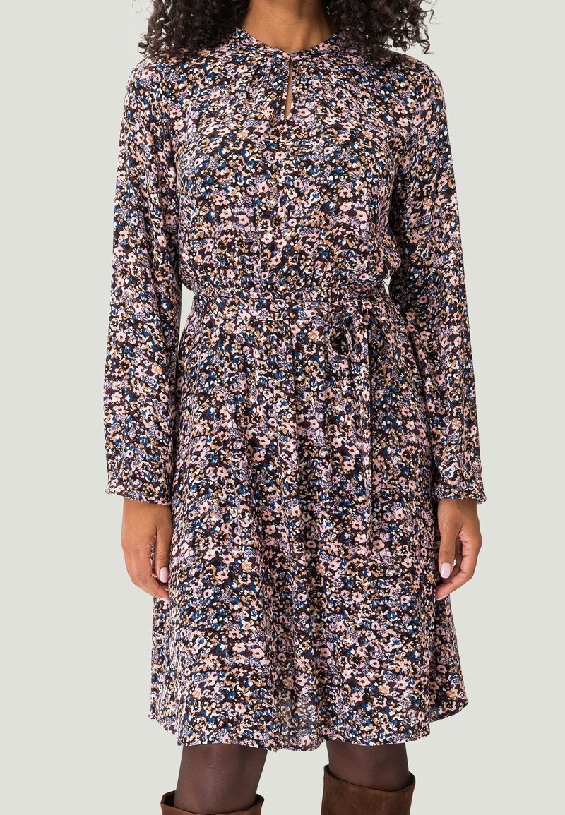 Femme portant une robe fleurie à manches longues, longueur genou, avec une ceinture à la taille et un décolleté en forme de trou de serrure, assortie à des bottes marron.
