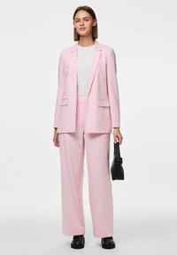 Blazer rose clair et pantalon assorti en tissu lisse, présentant une coupe classique avec des poches. Accompagné d'un haut blanc et d'un sac à main noir.
