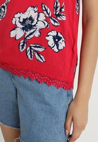 Blusa floral roja con flores azul marino, que presenta una tela texturizada y un dobladillo de encaje. Combinada con pantalones cortos de mezclilla azul claro.