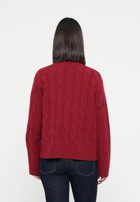 Rød kabelstrikket genser med ribbestrikket kant og vide ermer, som viser et teksturert mønster med et uformelt kort design.