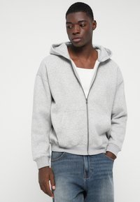 Jauns vīrietis, tērpts gaiši pelēkā pelēkā kapučjakā, baltā apakškreklā un zilos džinsos, stāv ar vienu roku kabatā pret vienkāršu fona.