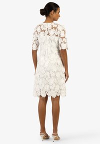 Weißes Spitzenkleid mit floralem Muster, kurzen Ärmeln und einem Reißverschluss auf der Rückseite. Es hat einen gewellten Saum und ist knielang.