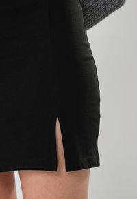 Gros plan d'une personne portant une jupe noire avec une petite fente à l'avant, montrant des jambes nues sur un fond neutre.