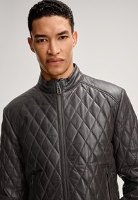 Mann mit kurzem lockigem Haar, der eine dunkelbraune gesteppte Lederjacke mit Frontreißverschluss und Taschen mit Reißverschluss trägt, neutraler Hintergrund.