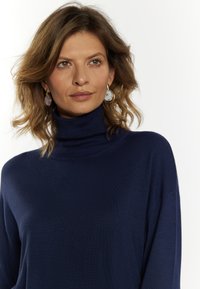 Dunkelblaues Turtleneck-Pullover aus weichem Strickstoff, mit lockerem Schnitt und geripptem Kragen. Das Model trägt runde Ohrringe.