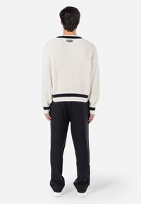 Ribbed white sweater with a black stripe at the hem and cuffs, featuring a logo on the back; paired with black wide-leg trousers.