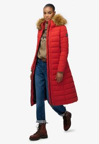 Rød, lang polstret frakke med en hætte i pels, der har lynlås lukning og et quiltet mønster. Kombinationen med blå jeans og brune snøresko.