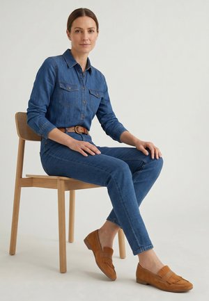 Kvinde iført denimskjorte og jeans med brunt bælte og sko, siddende på en lys træstol mod en ensfarvet baggrund.