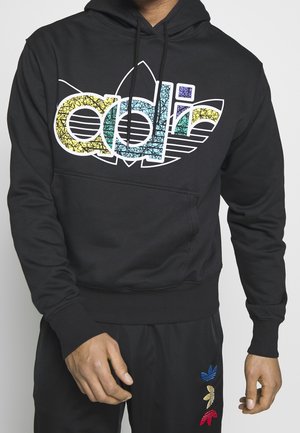Schwarzer Hoodie mit mehrfarbigem Adidas-Logo auf der Brust, kombiniert mit schwarzen Hosen mit drei kleinen Adidas-Logos in Blau, Gelb und Rot.