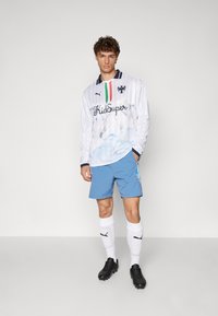 Weißes Langarmhemd mit blauem Marmor-Muster, "KidSuper"-Logo, marineblauer Kragen; kombiniert mit blauen Shorts und weißen Socken mit schwarzen Akzenten.