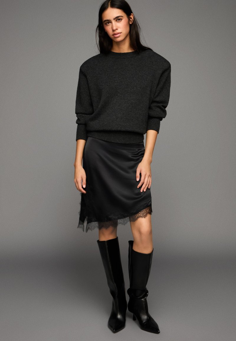 Čierny pletený sveter s nadrozmerným strihom, v páre s čiernou saténovou sukňou s čipkovaným lemom. Modelka má na sebe čierne vysoké čižmy na podpätku po kolená.