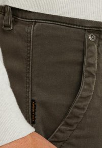 Pantalons en coton vert foncé avec une texture lisse, présentant des détails de couture subtils, un design de poche classique et une étiquette de marque.