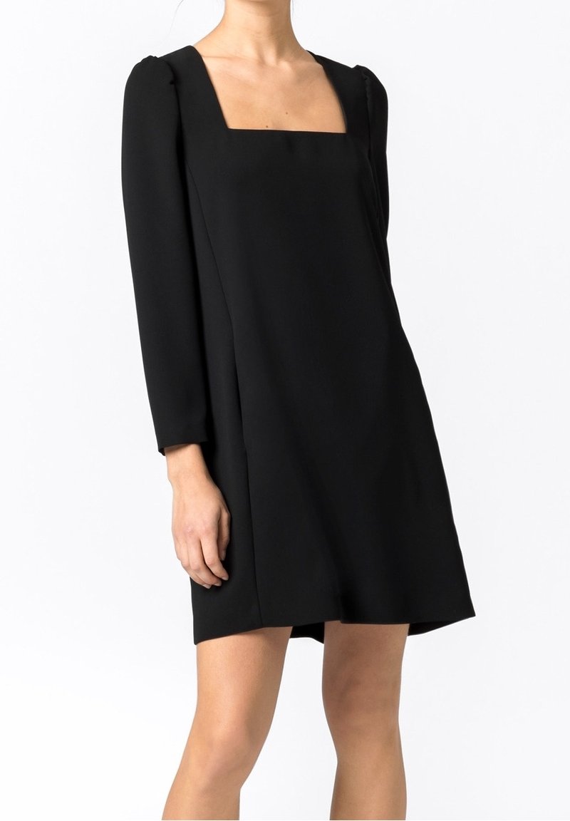 Femme portant une robe noire à manches longues avec un col carré, longueur au-dessus du genou, debout devant un fond blanc uni.