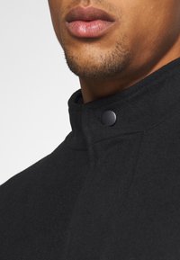 Černý vlněný kabát s vysokým, strukturovaným límcem a zapínáním na patentky. Textura je hladká s jemným matným povrchem.