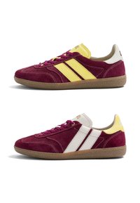 Paire de baskets en daim bordeaux avec semelles en gomme, lacets violets ; une chaussure a des rayures jaunes et un talon, l’autre des rayures blanches et un talon jaune.