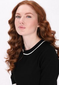 Pulover negru cu un tiv alb la guler, având o textură moale. Modelul are părul lung, ondulat și roșu și o expresie neutră. Fundal luminos.