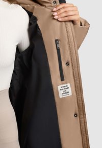 Barna pufidzseki fekete belsővel, oldalsó cipzáras zsebbekkel, és a "Egy meleg ölelés, ami szövetből készült" felirattal. Simább textúrával.