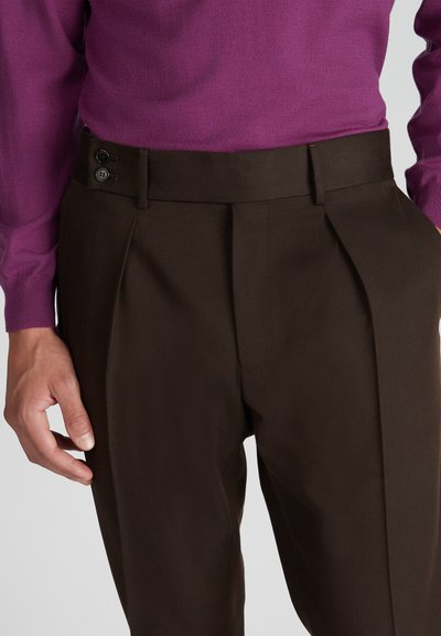 Pantaloni marroni a pieghe realizzati in tessuto testurizzato, dotati di due bottoni laterali in vita, abbinati a un maglione viola.