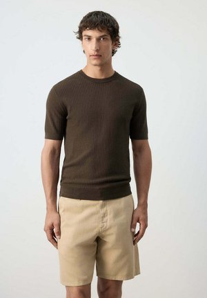 Homme portant un haut texturé à manches courtes marron foncé et un short beige, debout devant un fond clair uni.