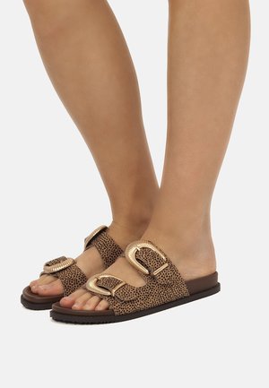 Pieds portant des sandales à enfiler marron avec des brides imprimées léopard et de grandes boucles dorées sur fond blanc.