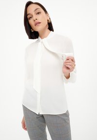 Blusa blanca y ligera con cuello puntiagudo y detalle de lazo en el cuello. Mangas largas con puños abotonados. Textura suave.
