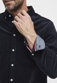Svart kordfløyelskjorte med krage med knapper; kontrasterende denimmansjett med røde og hvite striper; har hvite knapper og en brystlomme.