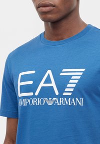 Άνδρας που φοράει μπλε μπλουζάκι με μεγάλο λευκό λογότυπο EA7 Emporio Armani στο μπροστινό μέρος, ορατό από τον λαιμό έως το μέσο του κορμού.