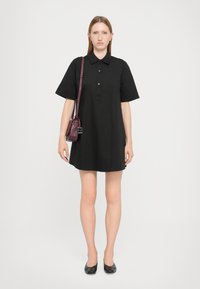 Rochie polo neagră cu mâneci scurte, cu închidere cu butoane, realizată dintr-un material neted. Asortată cu o geantă mică burgundă și pantofi negri.