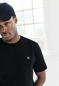 Must have lühikese varrukaga must t-särk, mille vasakul rinnal on väike valge logo; kangas näeb välja sile; kantud sobiva musta mütsiga.