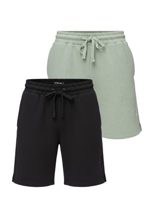 Man met baard die een wit grafisch T-shirt, zwarte korte broek, witte sneakers en zwarte polsbanden draagt, staande tegen een effen achtergrond.