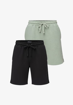 Mann mit Bart, der ein weißes grafisches T-Shirt, schwarze Shorts, weiße Sneakers und schwarze Handgelenkbänder trägt und vor einem schlichten Hintergrund steht.